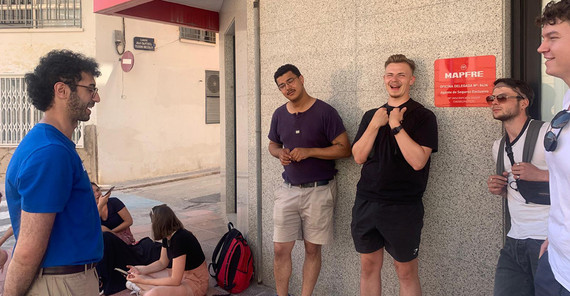 Eine Gruppe von Menschen steht, teils an einer Hauswand lehnend oder auf dem Boden sitzend, im Außenbereich. Sie wirken in ein unterhaltsames Gespräch verwickelt.
