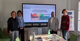 Vier Personen posieren für ein Gruppenfoto vor einem großen Bildschirm.