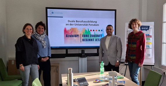 Vier Personen posieren für ein Gruppenfoto vor einem großen Bildschirm.