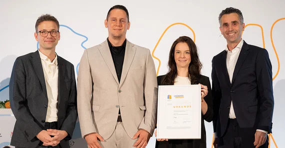 Vier Personen posieren für ein Foto. Eine hält eine große Urkunde.