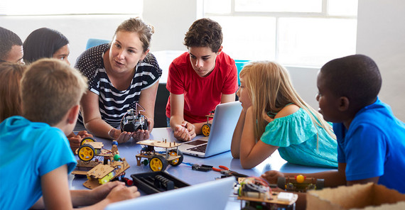 Eine Gruppe von jungen Schüller*innen umringt eine Lehrperson, die ihnen etwas erklärt.