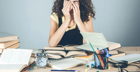Gestresste Studentin mit Büchern um sich herum auf dem Schreibtisch.