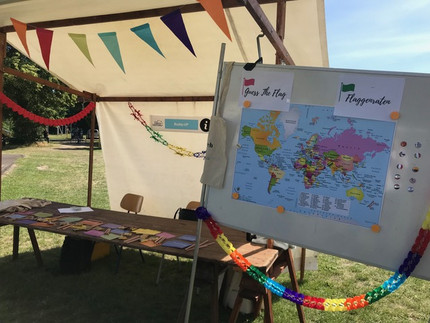 Ein Stand mit Tisch und Stühlen, der mit bunten Wimpelketten und Flyern dekoriert ist. Daneben eine Magnetwand, auf der eine Weltkarte angebracht ist. Neben der Karte sind 10 Magneten mit unterschiedlichen Flaggen bedruckt, über der Weltkarte steht "Guess the Flag" und "Flaggenraten".