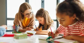 Eine Lehrerin hilft einer Schülerin, daneben sitzt eine zweite Schülerin