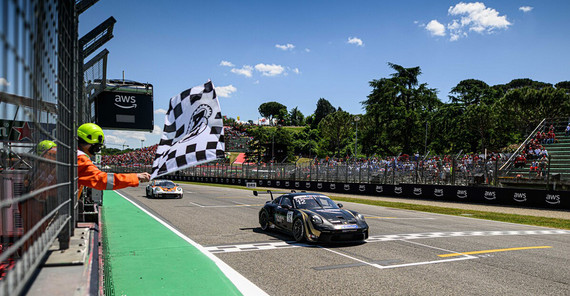 Flynt Schuring bei der Zieleinfahrt in Imola, wo er die schnellste Rennrunde gefahren ist Ein schwarzer Porsche 911 mit goldenen Details fährt über die Ziellinie. Links schwenkt jemand mit Helm und Kopfhörern eine schwarz-weiß-karierte Zielflagge. Im Hintergrund sieht man volle Zuschauerränge und Bäume. Der Himmel ist blau mit einzelnen, weißen Wolken.