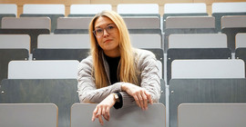 Frau sitzt im Hörsaal in Sizreihen und blickt in die Kamera.