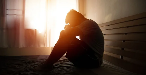 Man sitting on bed with head on knees. The picture is from AdobeStock/kwanchaift.