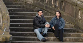 Zwei Personen sitzen auf einer Treppe und schauen in die Kamera.