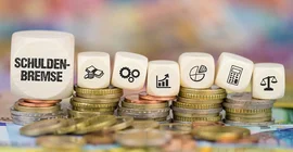 Symbolbild: Auf mehrern Stapel Hartgeld liegen diverse Spielwürfel. Auf ihnen sind u.a. Symbole für Berechnungen und Statistiken zu sehen. Auf dem größten Würfel ganz links steht "Schuldenbremse".