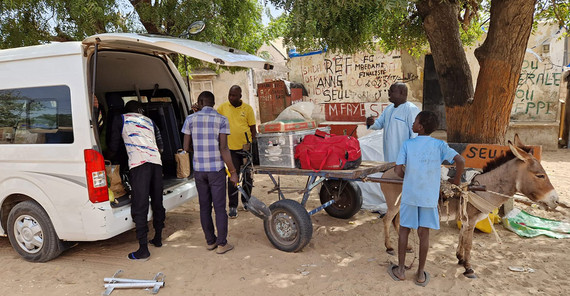 Fünf Personen stehen vor einem weißen Transporter und verladen technische Ausrüstung auf einen Esel der einen Wagen zieht.