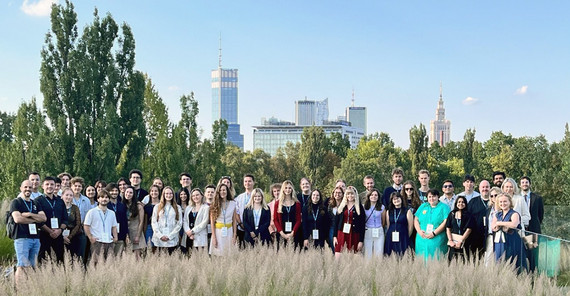 Studentische Vertretende, die sich auf einer Wiese zu einem Gruppenbild zusammengefunden haben. Im Hintergrund die Skyline Warschaus.