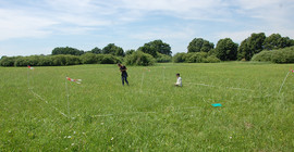 Bild von einer Wiese, auf der ein Stück mit Flatterband abgesperrt ist, darauf zwei junge Menschen