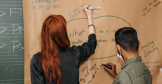 Eine Frau und ein Mann schreiben mit Stiften etwas auf ein großes Blatt Papier, das an eine Tafel gepinnt ist.