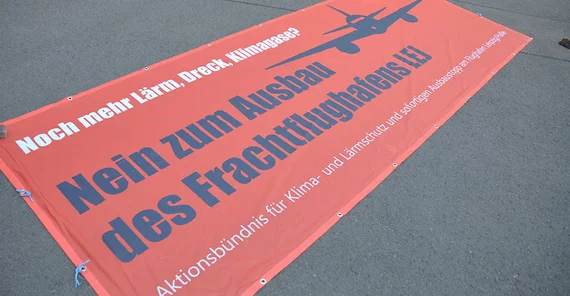 Rotes Protestbanner liegt auf dem Boden mit den Worten "Noch mehr Lärm, Dreck und Klimagase? Nein zum Ausbau des Frachtflughafens (E) Aktionsbündnis für Klima- und Lärmschutz und sofortigen Ausbaustopp am Flughafen Leizig/Halle"
