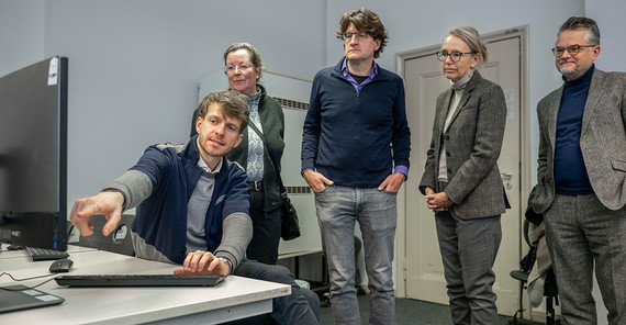 Eine Gruppe von Menschen steht in einem Raum vor einem Computer, während einer etwas erläutert.