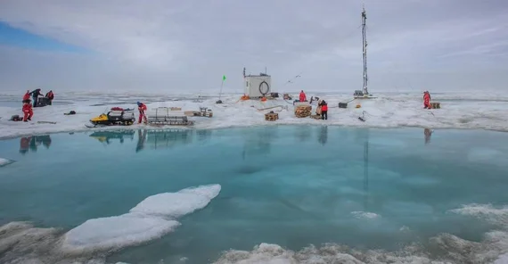 Eistümpel vor einer kleinen Forschungsstation in Polarregion. Mehrere Menschen in signalroten Overalls arbeiten mit Forschungsequipment.