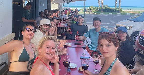Die Gruppe von Lehrenden und Studierenden sitzt über zwei Tische verteilt im Außenbereich einer Gaststätte. Alle haben Getränke vor sich und blicken in die Kamera. Im Hintergrund sind Meer und Horizont zu erkennen.