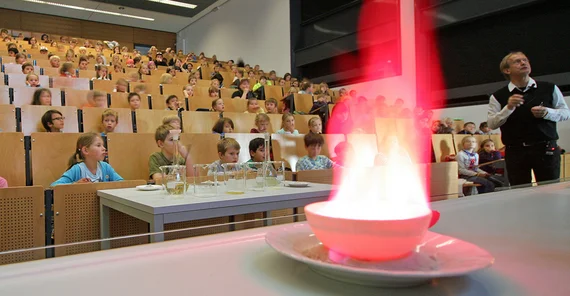 Eine rote, helle Flamme kommt aus einem Porzellangefäß. Im Hintergrund erklärt Prof. Holdt etwas dazu, Kinder im Hörsaal schauen zu.