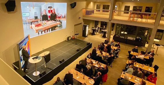 Blick in eine Veranstaltung mit vielen Menschen in einem Saal, zwei Personen sind auf einer Leinwand zugeschaltet