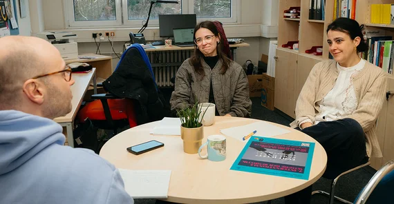 Drei Personen sitzen an einem runden Tisch im Gespräch.