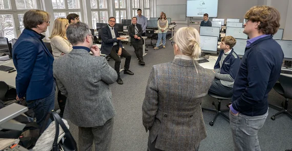 Eine Gruppe von Menschen steht in einem Raum mit Computern im Kreis und unterhält sich.