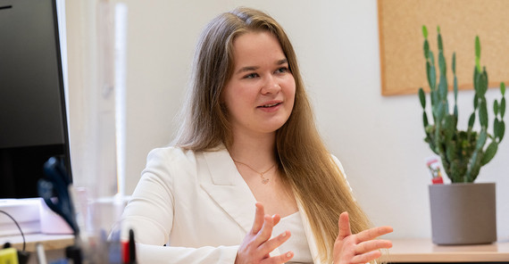 Eine junge Frau ein weiß erklärt etwas, im Hintergrund stehen ein Kaktus und Büromaterialien.