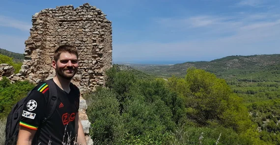 Eine Person im Vordergrund lacht in die Kamera. Hinter ihr Ruinen und grüne Vegetation. Die Person und die Ruine befinden sich auf einem Berg.